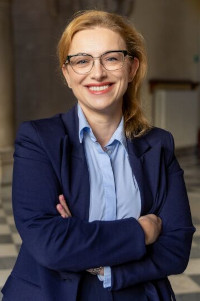 Uśmiechnięta kobieta zjasnymi włosami w okularach. Ubrana w niebieską koszulową bluzkę i granatową marynarkę. Sylwetka od pasa w górę. Trzyma ręce na wysokości pasa założone jedna na drugą. W tle hol kamienny w pałacu Wielopolskich, na zdjęciu widać ścianę i posadzkę w czarno-bialy wzór.